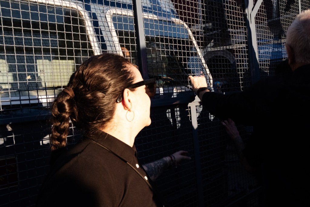 A woman with a braid and sunglasses looks through a metal mesh barrier at a police van; a man's hand rests on the fence beside her.