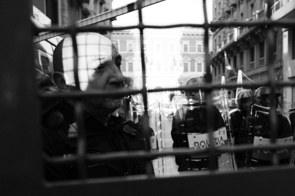 A police man with grey stubble looks through the grid of a barrier at riot police holding shields marked "POLIZIA".
