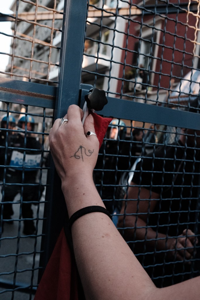 A hand with rings and a small tattoo holds a red-and-white cloth against a barrier knob; riot police in helmets stand on the other side of the mesh.