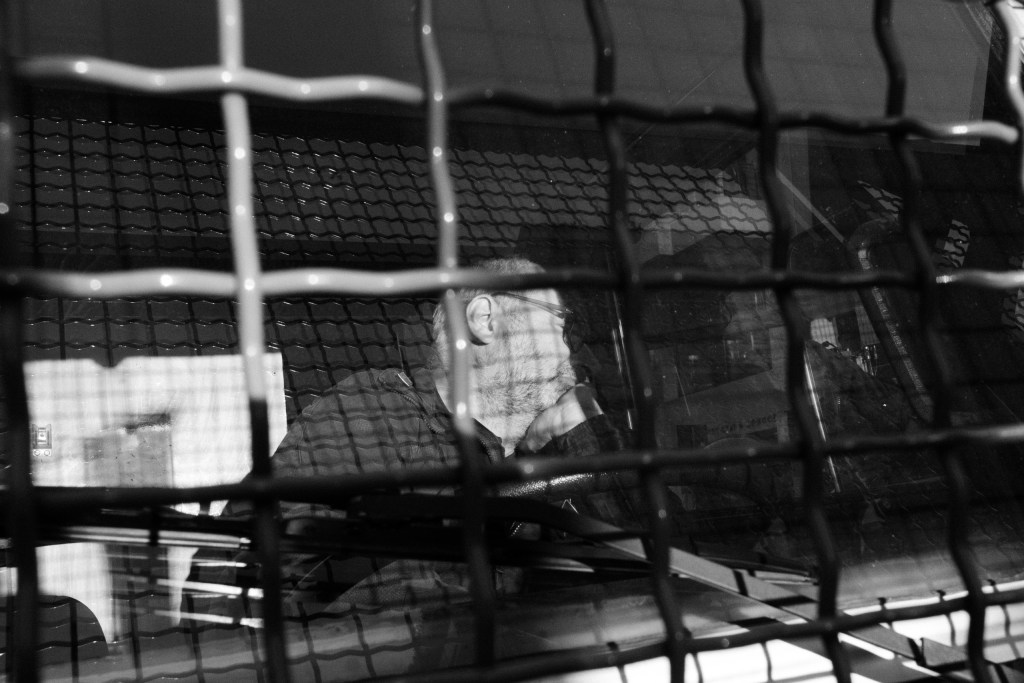 Seen through metal mesh and window glass layered with reflections, a bearded policeman with glasses sits with his hand at his chin.