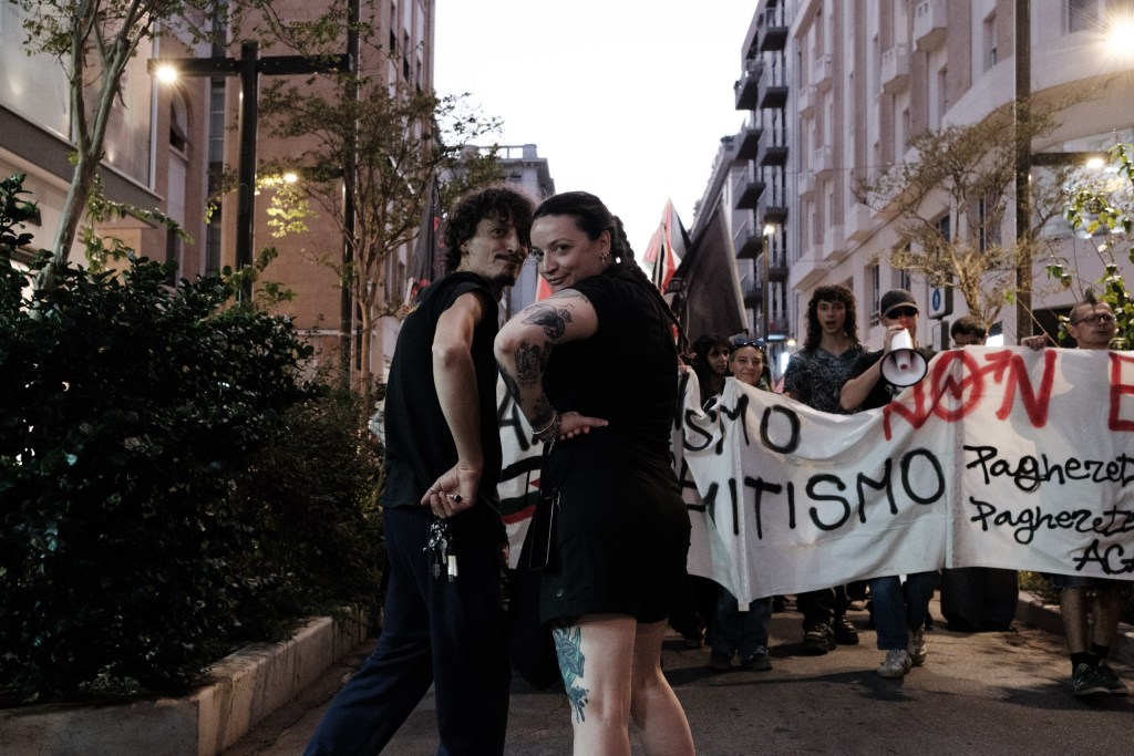 At dusk, two people look back toward the camera on a tree-lined street; behind them the march follows a white banner with sprayed lettering, and a person holds a megaphone.