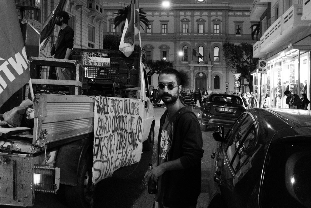 At night, a man with round sunglasses and a bottle in his hand looks at the camera in front of a flatbed truck carrying a sound system and a banner that begins "Israele terrorista, Italia complice".