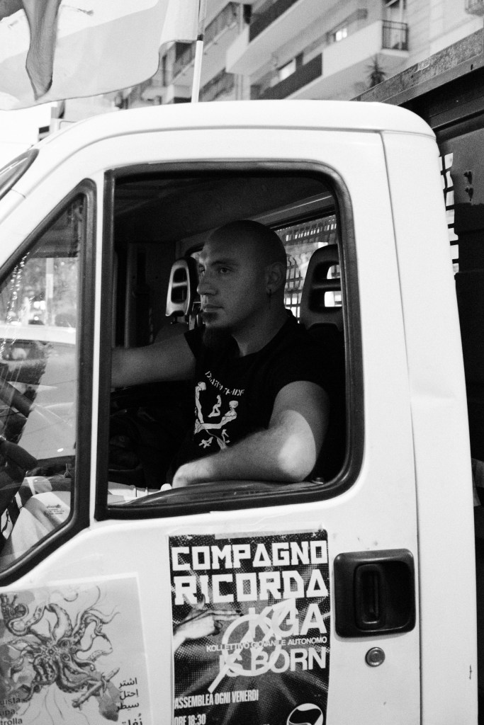 A bald man with a beard sits at the wheel of a white truck, seen through the open cab window; stickers cover the door below.