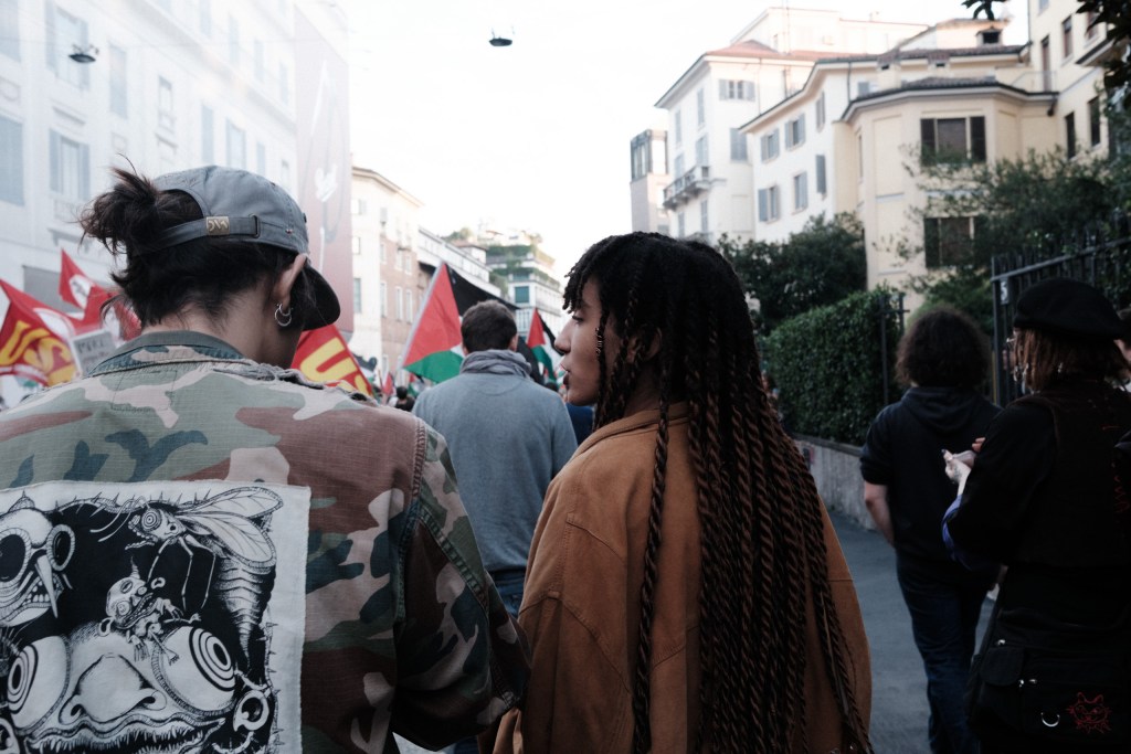 Two marchers seen from behind and in profile: one wears a camouflage jacket with a large illustrated insect patch, the other has long braided hair and looks to the side, with union and Palestinian flags behind them.