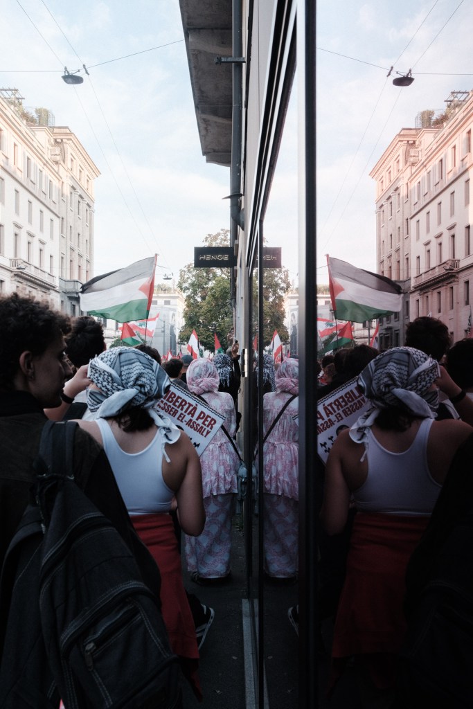 Marchers in keffiyeh headscarves walk away from the camera along a street of old apartment buildings, mirrored in a glass shopfront, with Palestinian and Lebanese flags ahead and a placard demanding freedom for a prisoner.