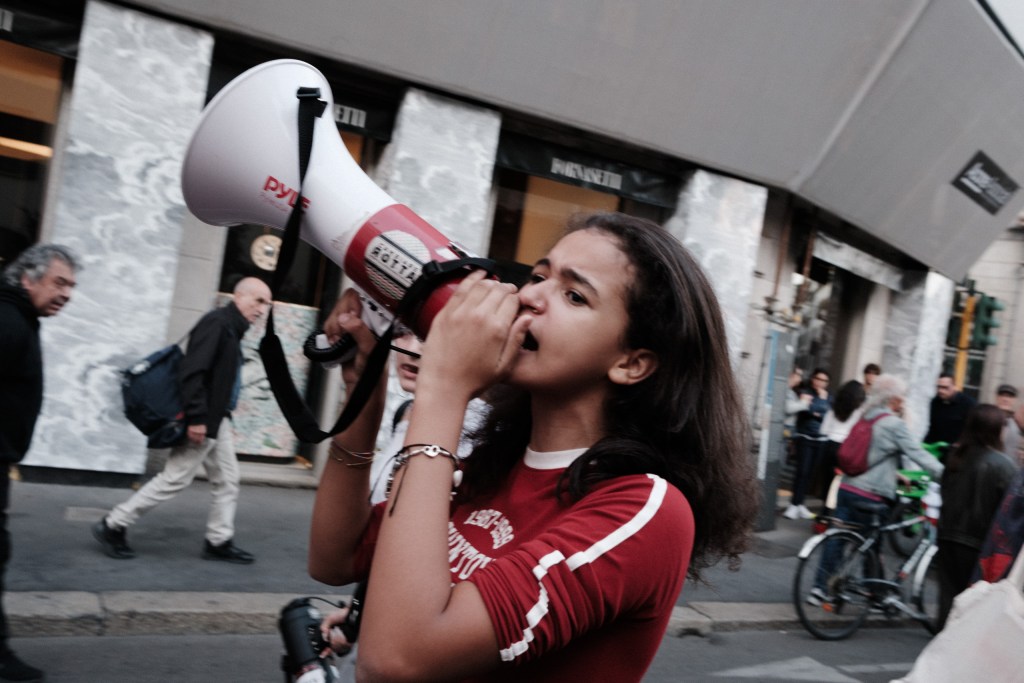 A young woman shouts into a megaphone while marching, with passersby and a parked bicycle behind her on the street.
