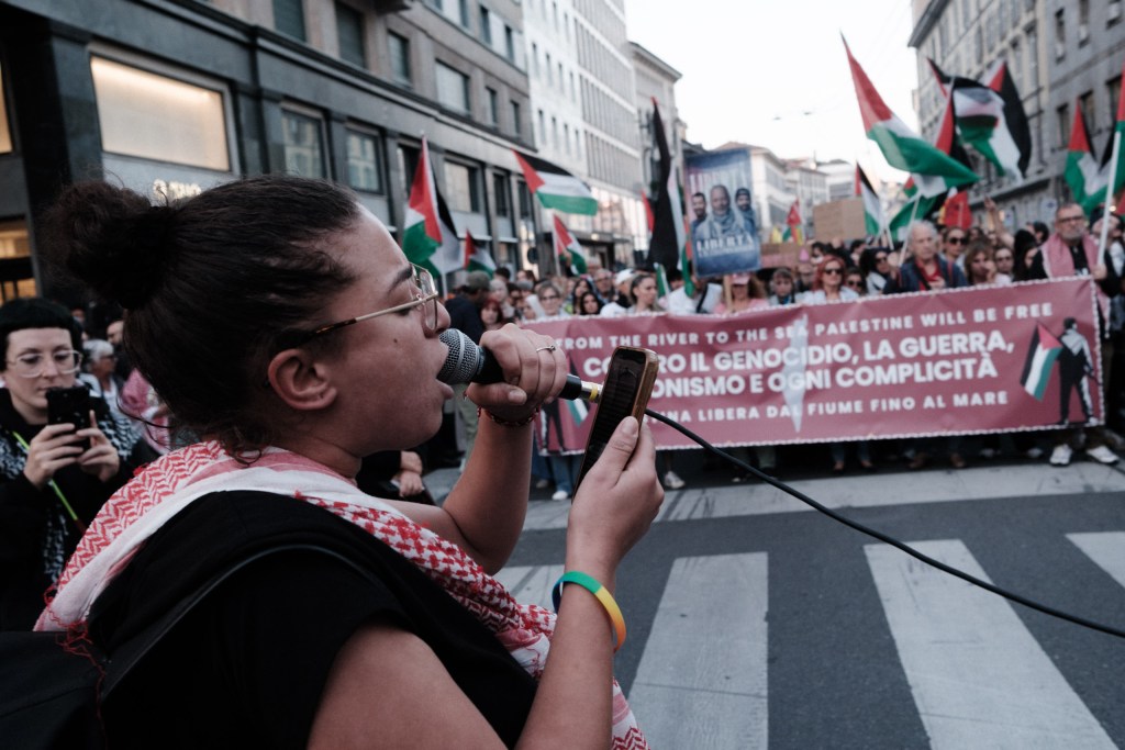 A woman in a keffiyeh speaks into a microphone connected to a phone, facing the lead banner and a crowd of marchers with Palestinian flags behind her.