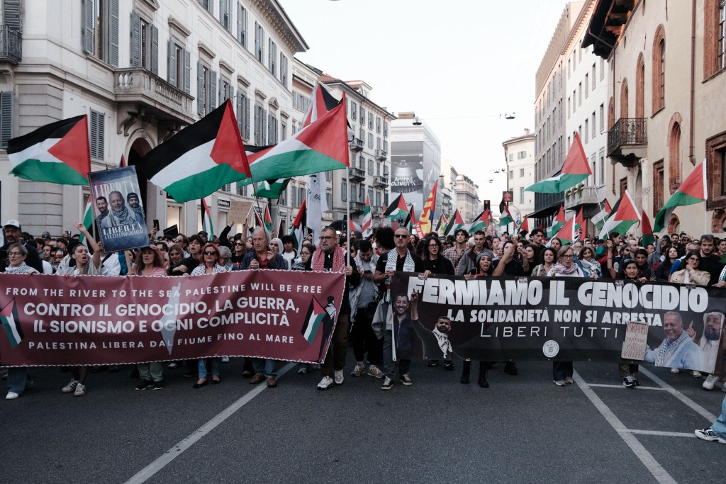 The head of the march seen from the street level: two large banners, one maroon and one black, held across the road beneath a mass of Palestinian flags.