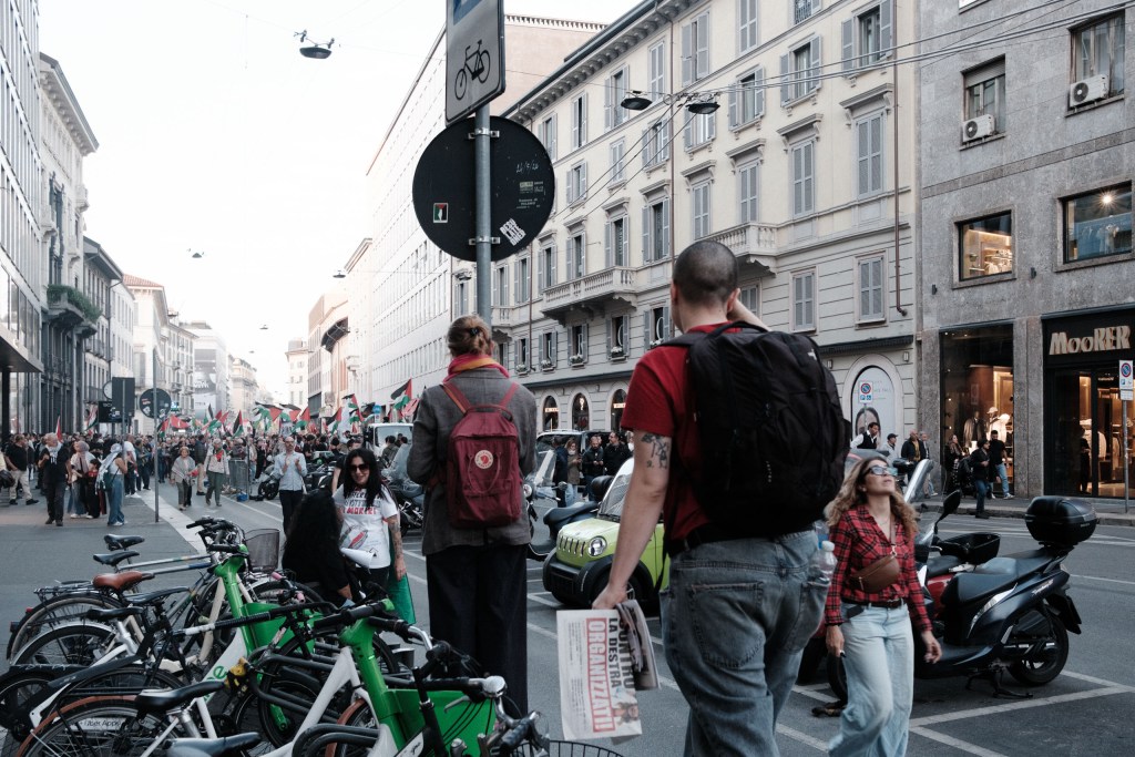 A street corner with a rack of shared bicycles and scooters in the foreground, as two onlookers seen from behind watch the march advance in the distance, one holding a folded newspaper.