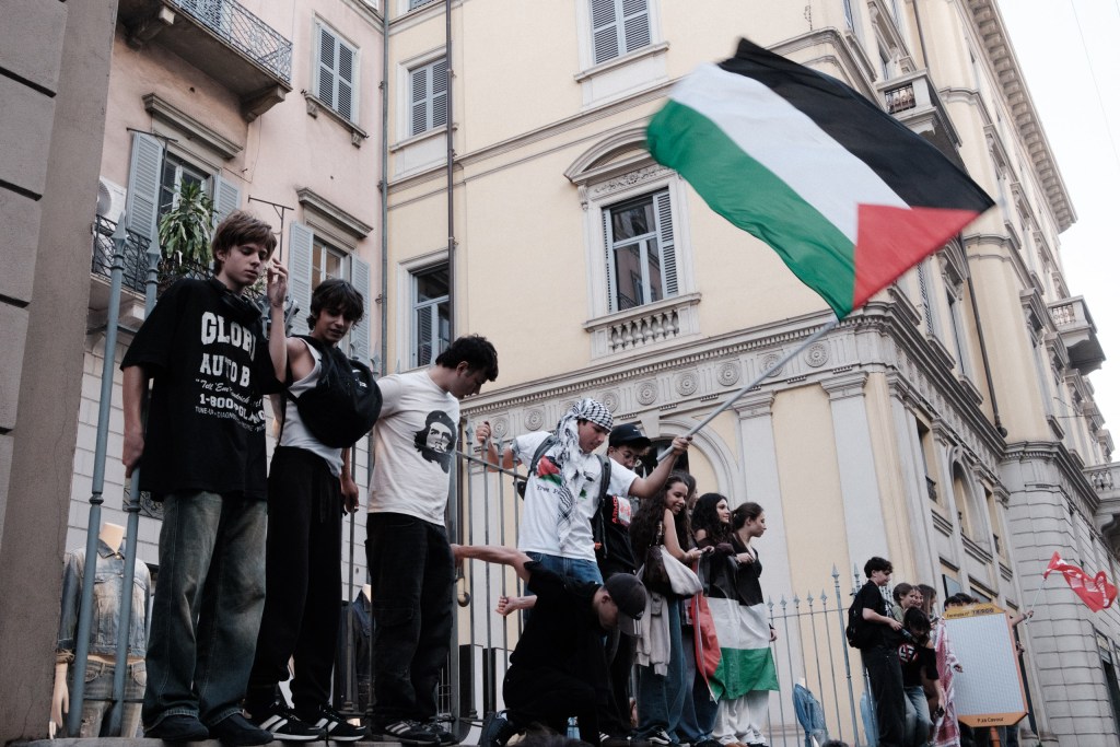 Teenagers stand along a wrought-iron railing above the march, one waving a Palestinian flag while others look down at the street, and a flag draped over a girl’s shoulders at right.