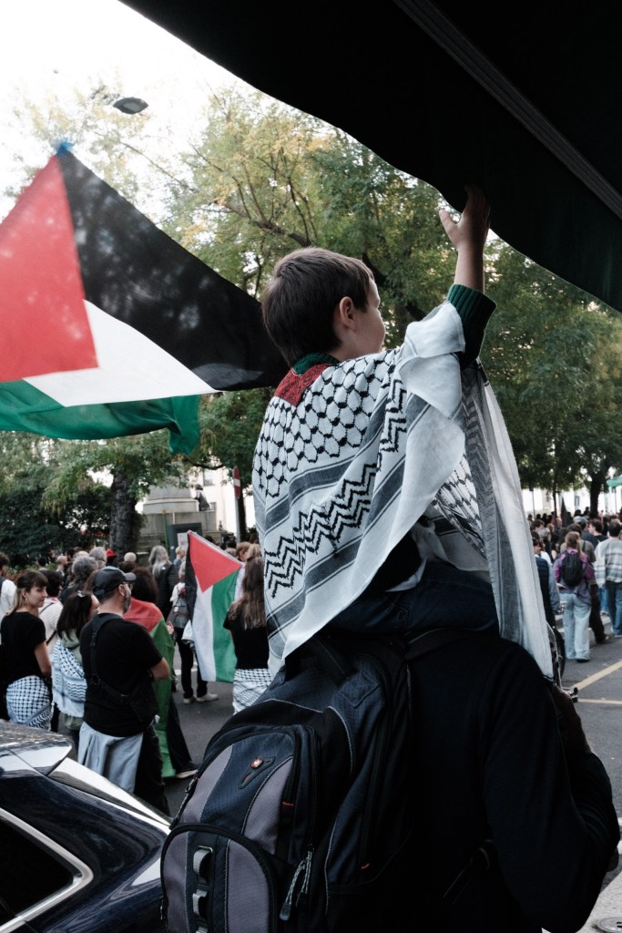A child wearing a keffiyeh sits on an adult’s shoulders and reaches up to touch a dark awning, with a Palestinian flag blurred beside them and the march behind.