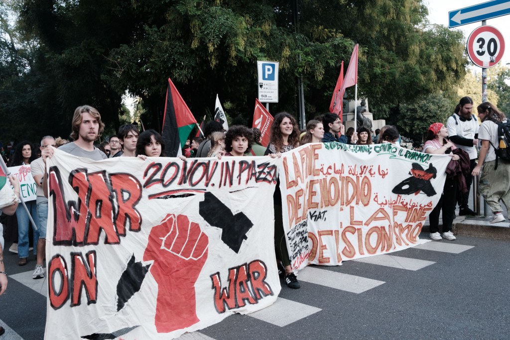 Two hand-painted banners held by young marchers: one reads “War on War” and “20 Nov. in piazza” with a red fist and a bomb, the other calls for the end of genocide and Zionism in Italian and Arabic.