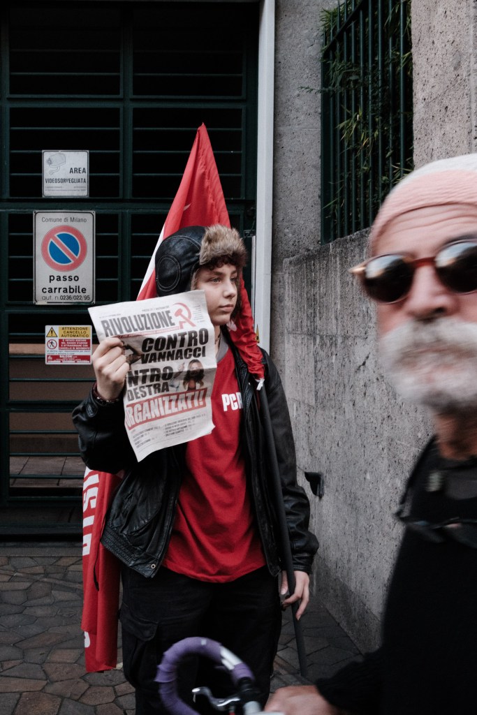 A teenager in a fur-lined cap and red shirt holds up a copy of the newspaper Rivoluzione, with a red flag behind, while a blurred bearded man in sunglasses passes in the foreground.