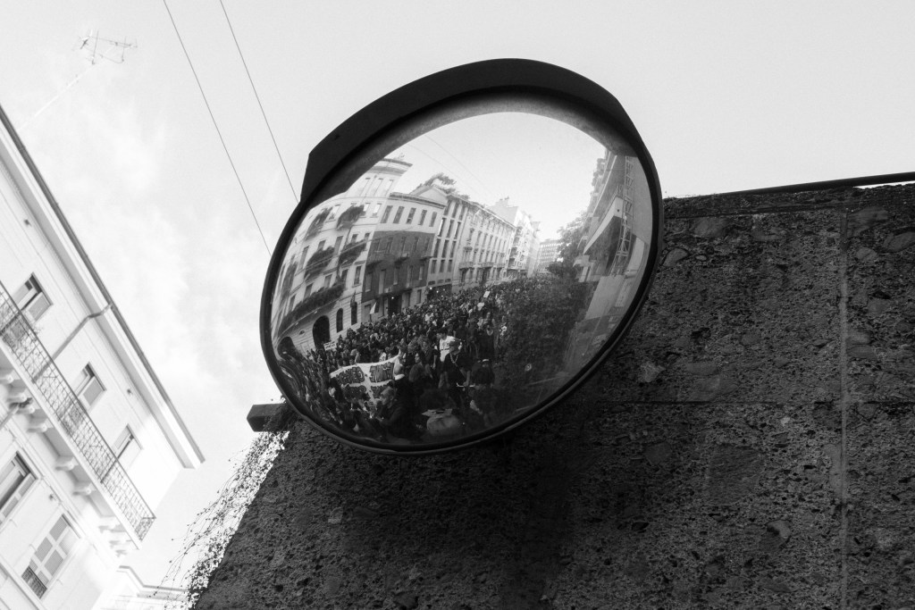 A convex traffic mirror mounted on a rough stone wall reflects the crowd filling the street, with a banner visible among the marchers and buildings curving along the edge of the glass.