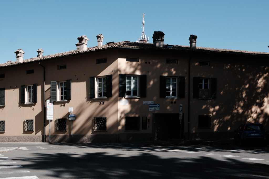 Two-storey ochre corner building with dark shutters, barred ground-floor windows and chimney pots, partly in tree shadow, with a radio mast above the roof and street signs below.