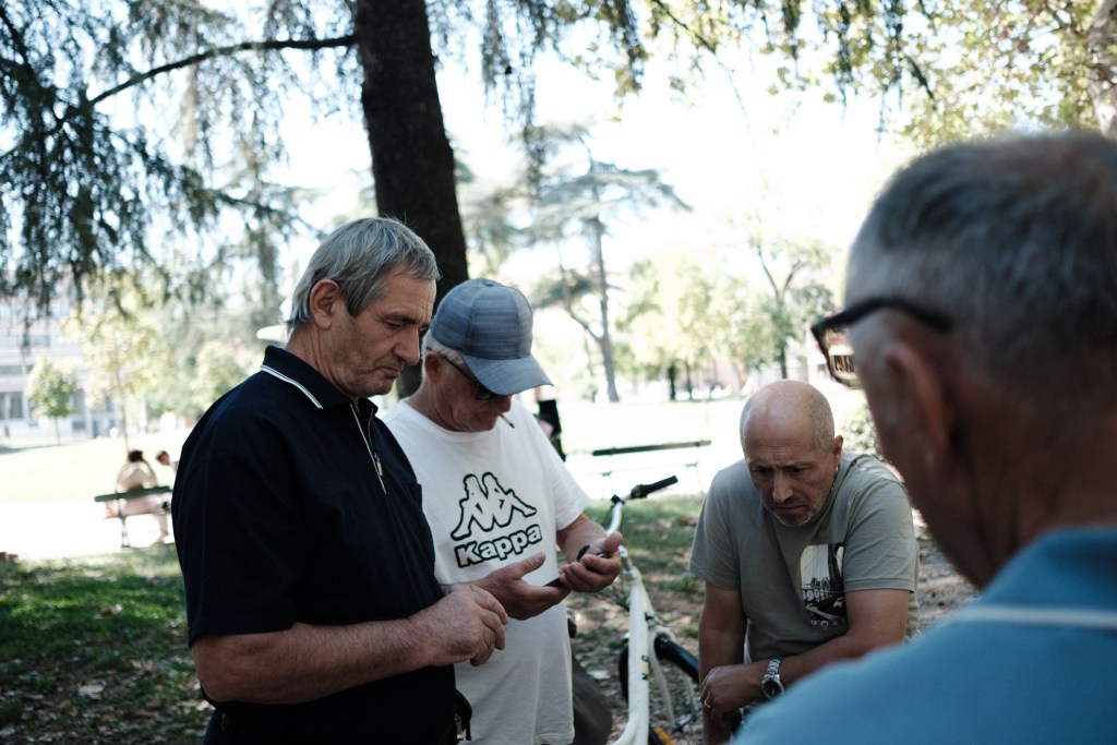 Four men stand close together in a park, heads lowered toward something held in hand, one with a cigarette in his mouth and a bicycle behind them.