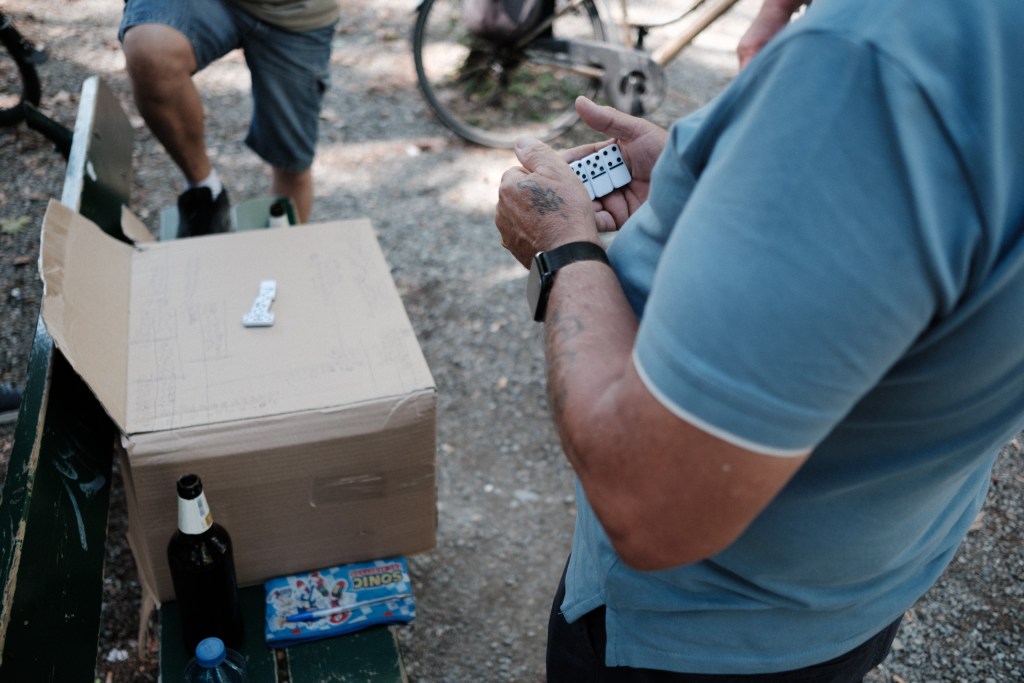 A man holds a handful of dominoes beside a cardboard box covered in handwritten scores, with a beer bottle and a child’s pencil case on the bench.