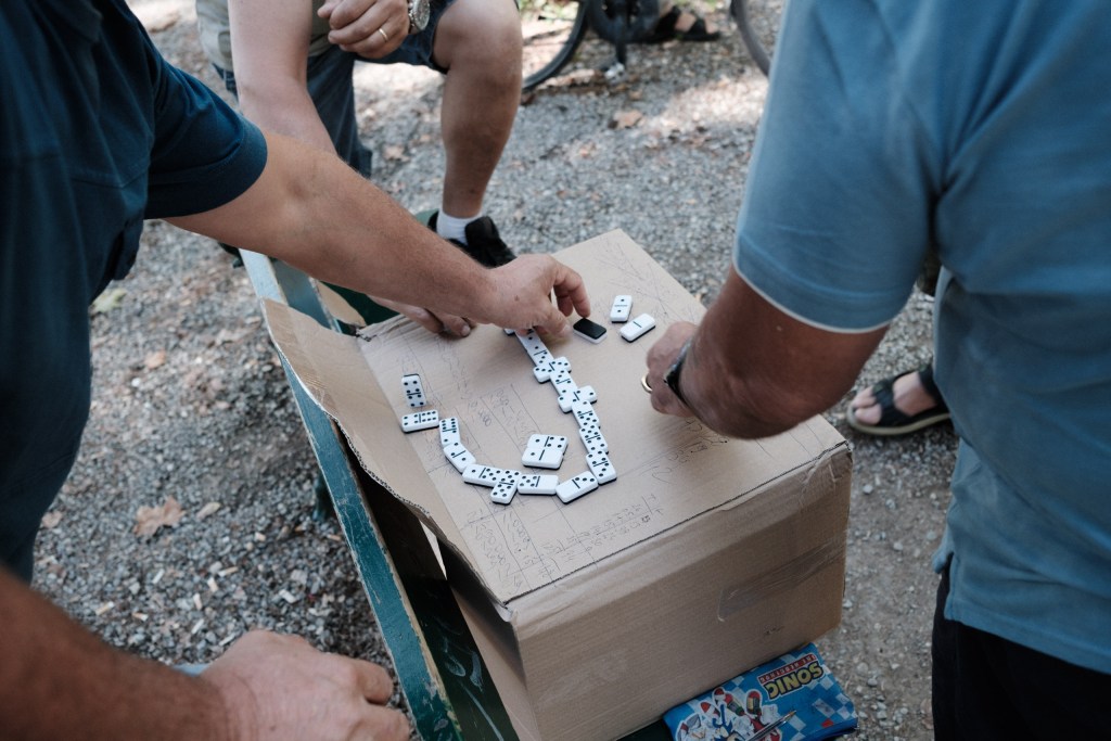 Hands reach over a cardboard box where dominoes form a winding line, with scores written across the cardboard and gravel on the ground below.