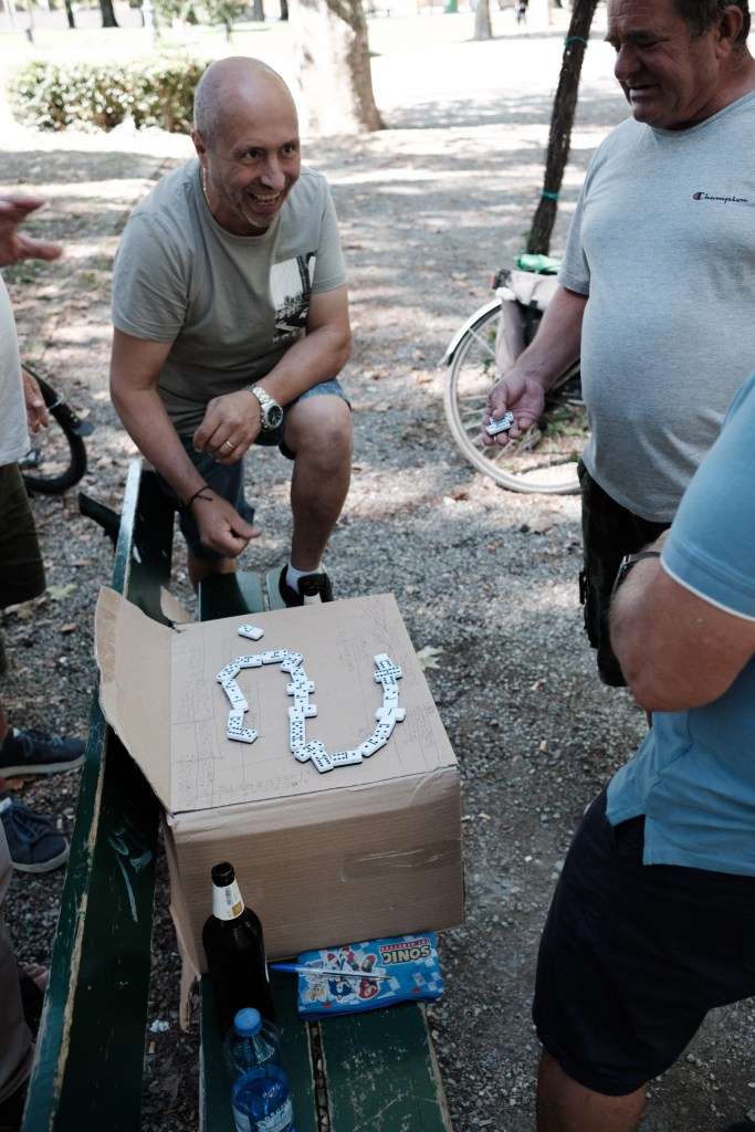 A smiling man crouches beside a cardboard box with dominoes laid in a loop while another man holds a tile, with a bottle, a water bottle and bicycles nearby.