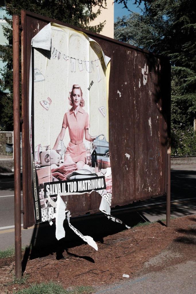 A rusted billboard holding a peeling poster of a woman in a pink 1950s-style dress surrounded by irons and laundry, with the words “Salva il tuo matrimonio” and “8 marzo 2026, Giornata internazionale delle donne”.