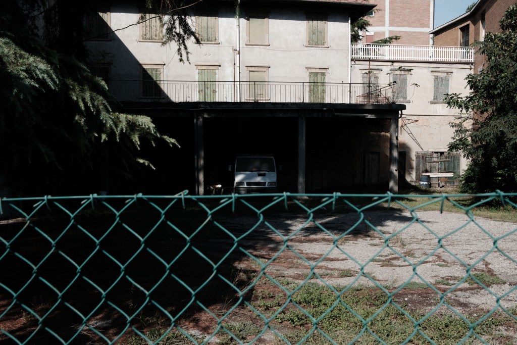 A white van parked in the shade of a covered area behind a green chain-link fence, in front of a weathered residential building with closed shutters.