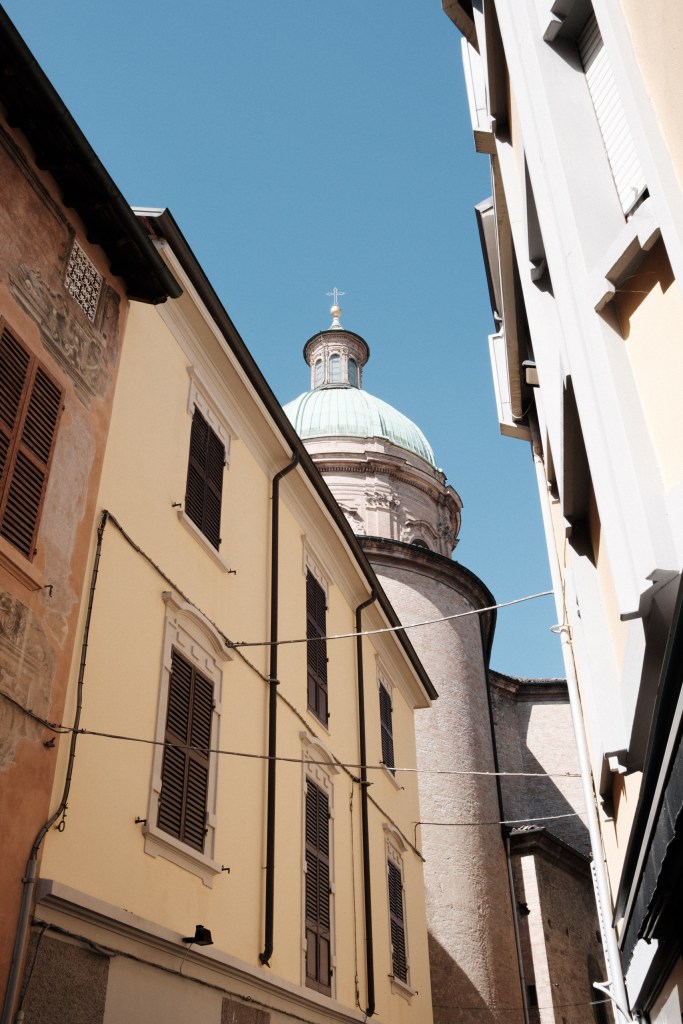 Looking up a narrow street at yellow and ochre façades with wooden shutters and overhead cables, with a green copper dome rising against a clear blue sky.