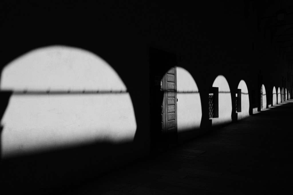 A row of arches along a portico, with curved patches of sunlight on the wall surrounded by deep shadow, a wooden door and small barred windows receding into the distance.