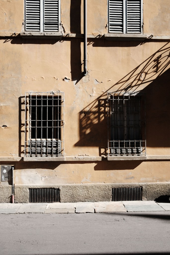A wall of peeling ochre plaster with two barred ground-floor windows and louvred shutters above, crossed by the diagonal shadow of an ornate ironwork balcony.